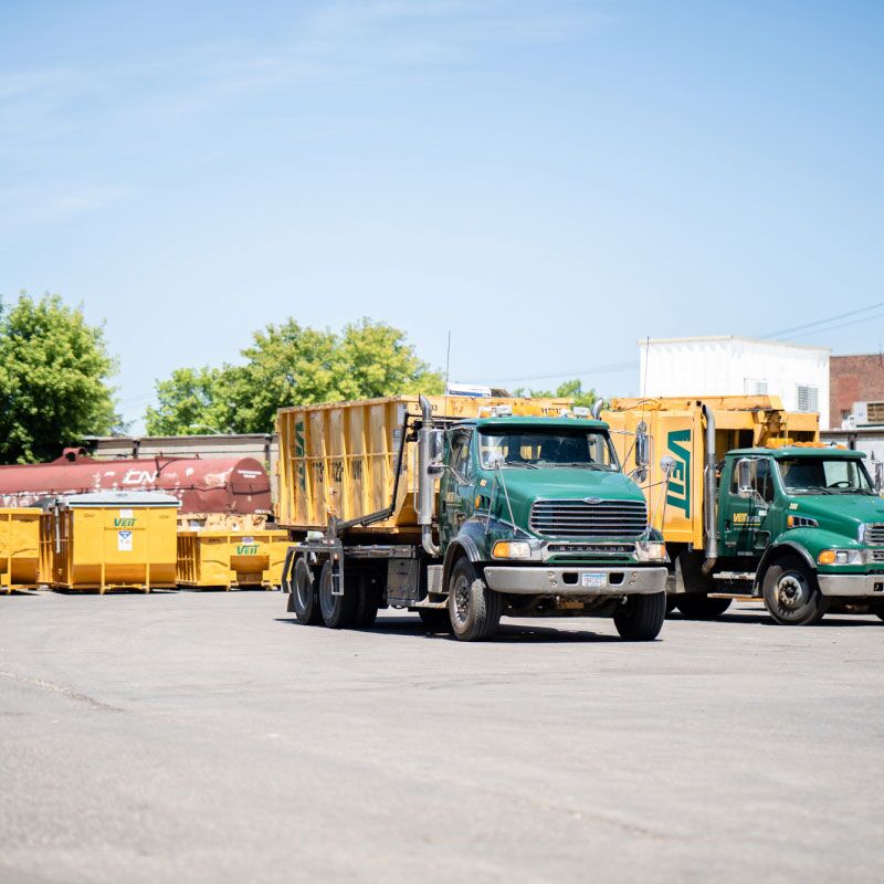 A parking lot featuring two Veit Disposal Mack dump trucks alongside roll-off dumpsters, emphasizing their industrial utility.